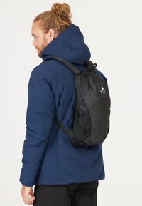 Black backpack with a textured design, mesh side pockets, and logo, worn over a blue hooded jacket. Features adjustable straps.