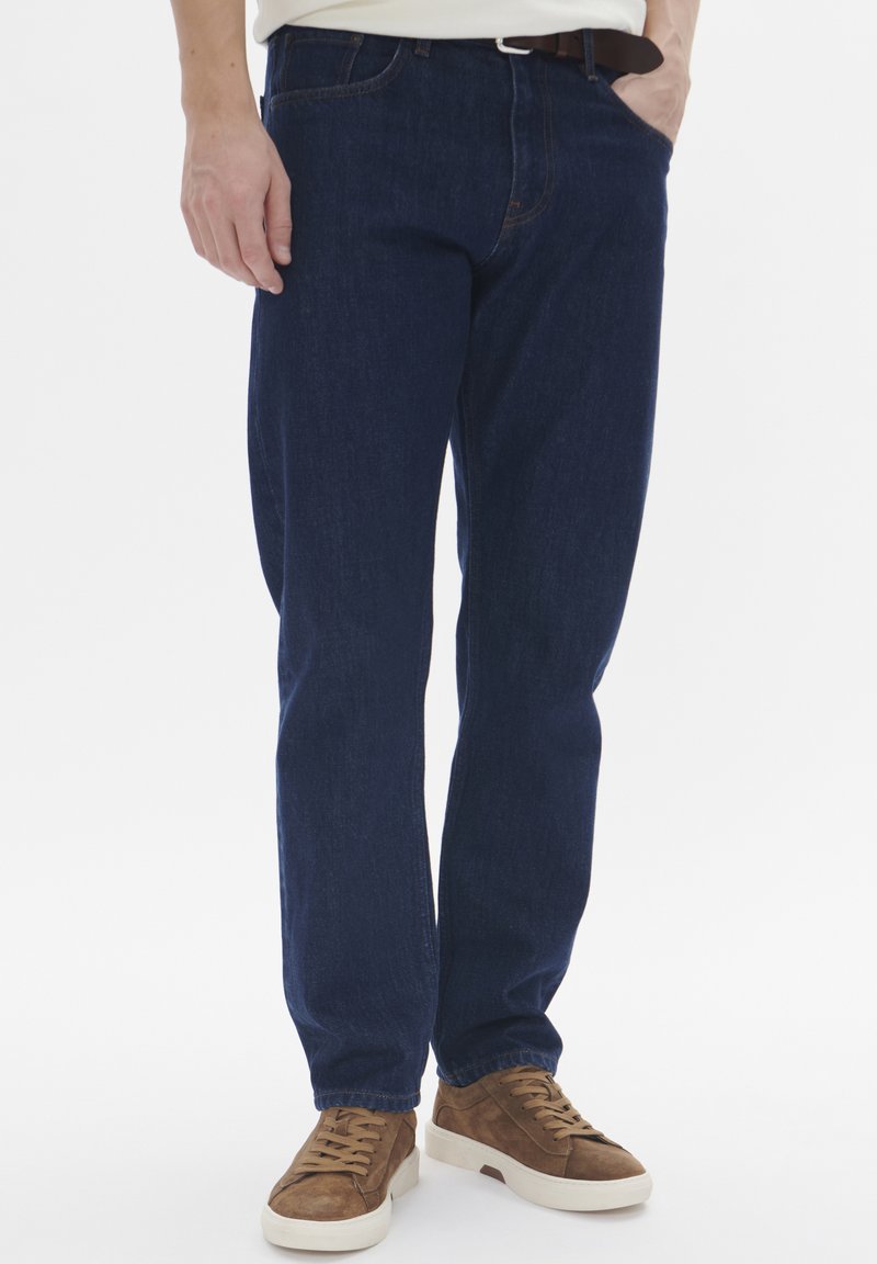 Temno modri jeans s ravnim krojem, ki vključujejo pet žepov in subtilne šive. Nosi se z rjavimi semiš supergami.
