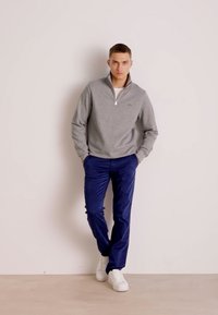 Felpa grigia con zip a quarto, pantaloni navy e sneaker bianchi. Il tessuto appare morbido, con una vestibilità rilassata e dettagli minimi.