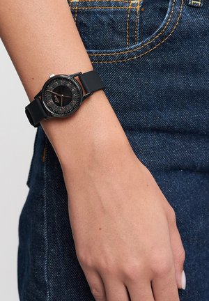 Schwarze Armbanduhr mit rundem Zifferblatt, strukturiertem schwarzem Ziffernblatt und goldenen Akzenten, montiert auf einem schwarzen Silikonarmband. Am Handgelenk getragen, vor einem Jeansstoff.