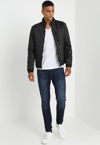 Svart quiltet jakke med standhals, glidelås, kombinert med en hvit V-hals skjorte og mørkeblå skinny jeans. Hvite joggesko fullfører antrekket.