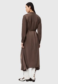 Vestito lungo marrone in satin con maniche lunghe, schiena plissettata, spacchi laterali e vita con cintura. Indossato con stivali al ginocchio bianchi e neri con motivo a fantasia.