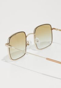 Firkantede solbriller med gullfarget metallramme, gradient brune glass og klare neseputer. Stengene har en slank design med roségull aksenter.