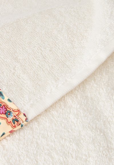 Tissu éponge blanc doux plié, avec un coin montrant une bordure beige à motifs avec des dessins floraux colorés.