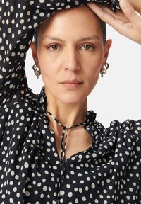 Blusa navy con lunares blancos, de tela texturada ligera, con un escote atado y mangas sueltas. Se ven pendientes de aro plateados.