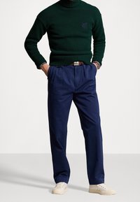 Mörkgrön polotröja med ribbad yta och blå logotyp, kombinerad med marinblå byxor, krämfärgade sneakers och ett brunt bälte.