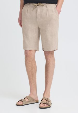 Hombre con pantalones cortos de lino beige con cintura ajustable y sandalias de ante taupe con doble correa, de pie con las manos en los bolsillos.