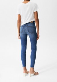 Person wearing blue skinny jeans, white short-sleeve t-shirt, and white heeled sandals, standing against a plain white background.