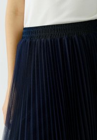 Jupe plissée bleu marine avec une large ceinture élastique texturée. Le tissu est transparent et léger, avec des drapés doux et fluides.