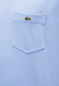 Ανοιχτό μπλε ύφασμα με μια ραμμένη τσέπη στο στήθος που διαθέτει ένα μικρό πράσινο λογότυπο κροκόδειλου κεντημένο κοντά στην πάνω άκρη της τσέπης.