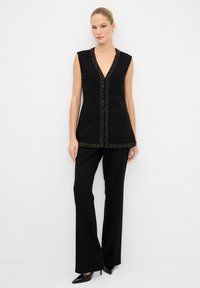 Chaleco negro sin mangas con ribete decorativo, dos bolsillos frontales, combinado con pantalones amplios negros y zapatos de tacón puntiagudos negros.