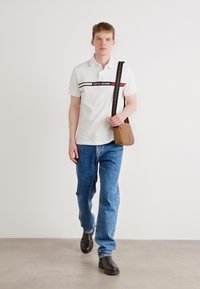 Hvit polo skjorte med en blå og rød horisontal stripe med "TOMMY HILFIGER" tekst, blå denim jeans og svarte slip-on sko. Brun skulderveske.