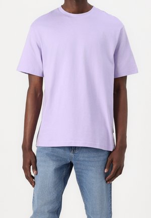 Mężczyzna ubrany w gładki jasnofioletowy t-shirt z krótkim rękawem i jasnoniebieskie dżinsy stojący na białym tle.