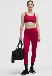 Žena v jasnoružovom športovom podprsenke a legínach drží čiernu športovú tašku a fľašu na vodu, má na nohách čierne tenisky a ponožky, stojí na jednoliatom pozadí.