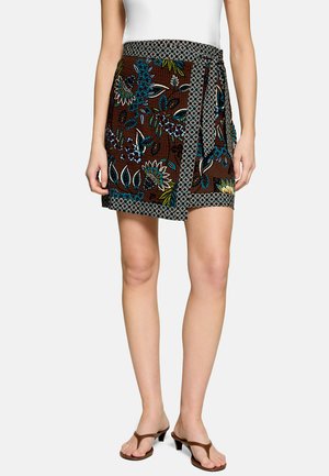 Femme portant une jupe portefeuille marron à fleurs avec une bordure à motifs, un haut blanc sans manches et des sandales marron à talons, debout devant un fond uni.