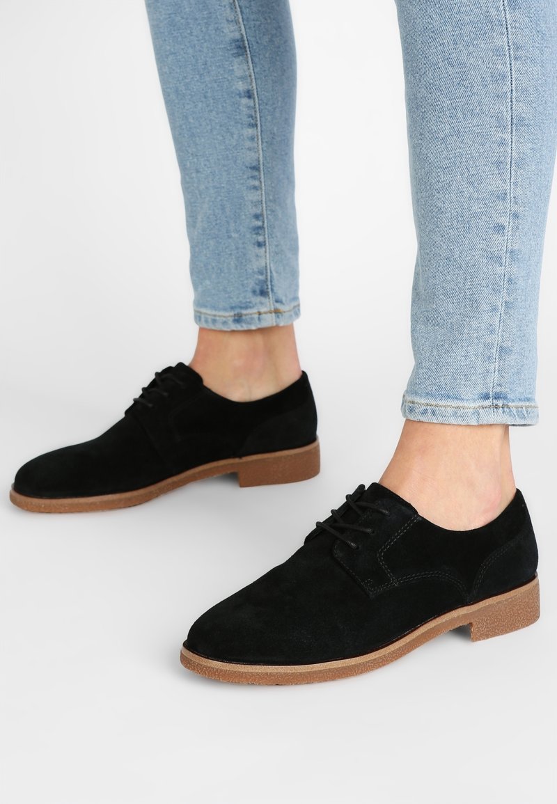 Personne portant un jean bleu clair et des chaussures montantes en daim noir avec des semelles marron sur un fond blanc.