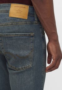 Крупен план на човек, носещ сини дънки от деним с кожена етикет Jack & Jones над джоб на гърба от дясната страна.