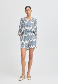 Rompere bianco con motivo paisley blu, scollo a v, maniche lunghe, vita cinchata e tasche. Abbinato a scarpe di colore chiaro.