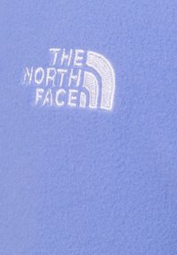 Jasnofioletowa tkanina polarowa z białym haftem logo "THE NORTH FACE". Miękka w dotyku, bez widocznych wzorów ani elementów metalowych.