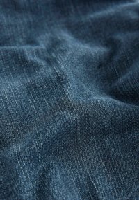 Tejido de mezclilla azul oscuro con una superficie texturizada, que muestra patrones de tejido visibles y un sutil brillo, dispuestos en suaves pliegues.