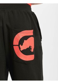 Sorte joggebukser med en stor rød grafikk av et neshorn på baklommen, med en strukturert lomme-design.