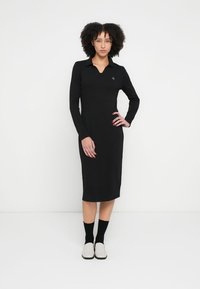 Schwarzes langärmliges Kleid mit Polokragen aus glattem Stoff. Knielang mit figurbetonter Silhouette und kleinem Logodetail auf der Brust.