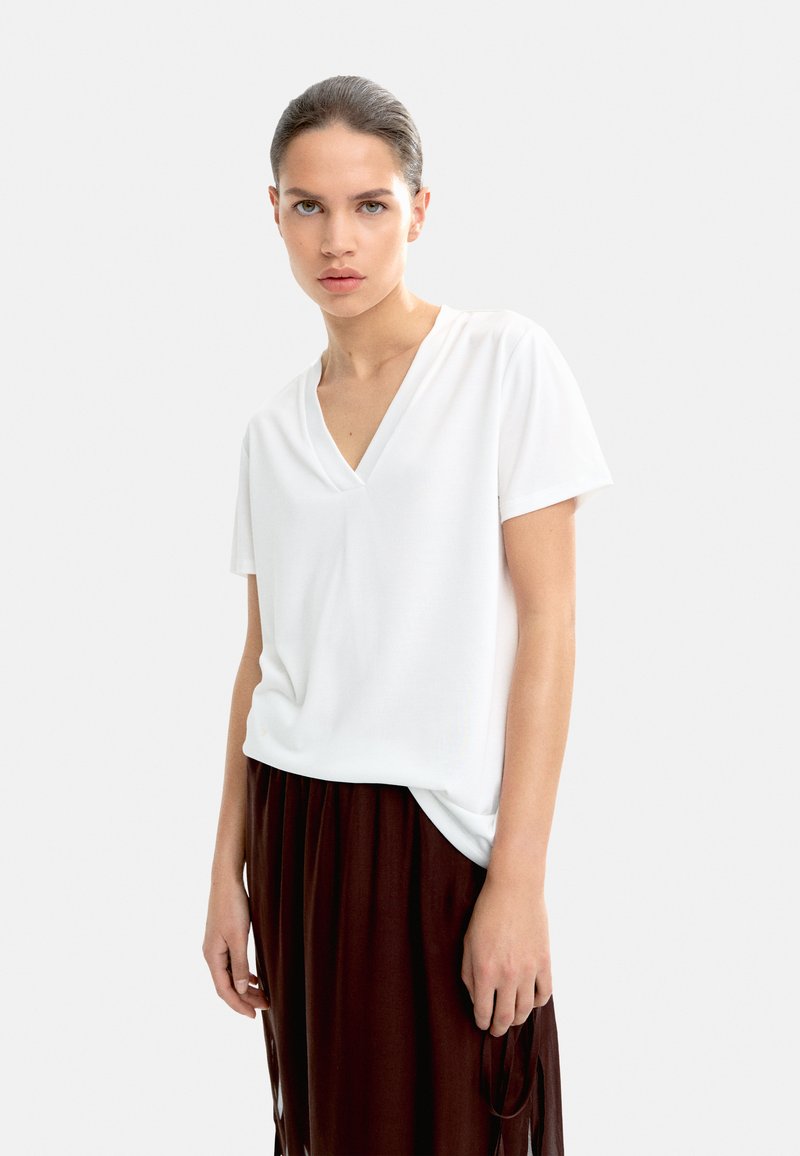 Tricou alb cu mânecă scurtă și decolteu în V, confecționat dintr-un material neted și ușor, asortat cu o fustă maro vaporoasă ce prezintă accente de franjuri.