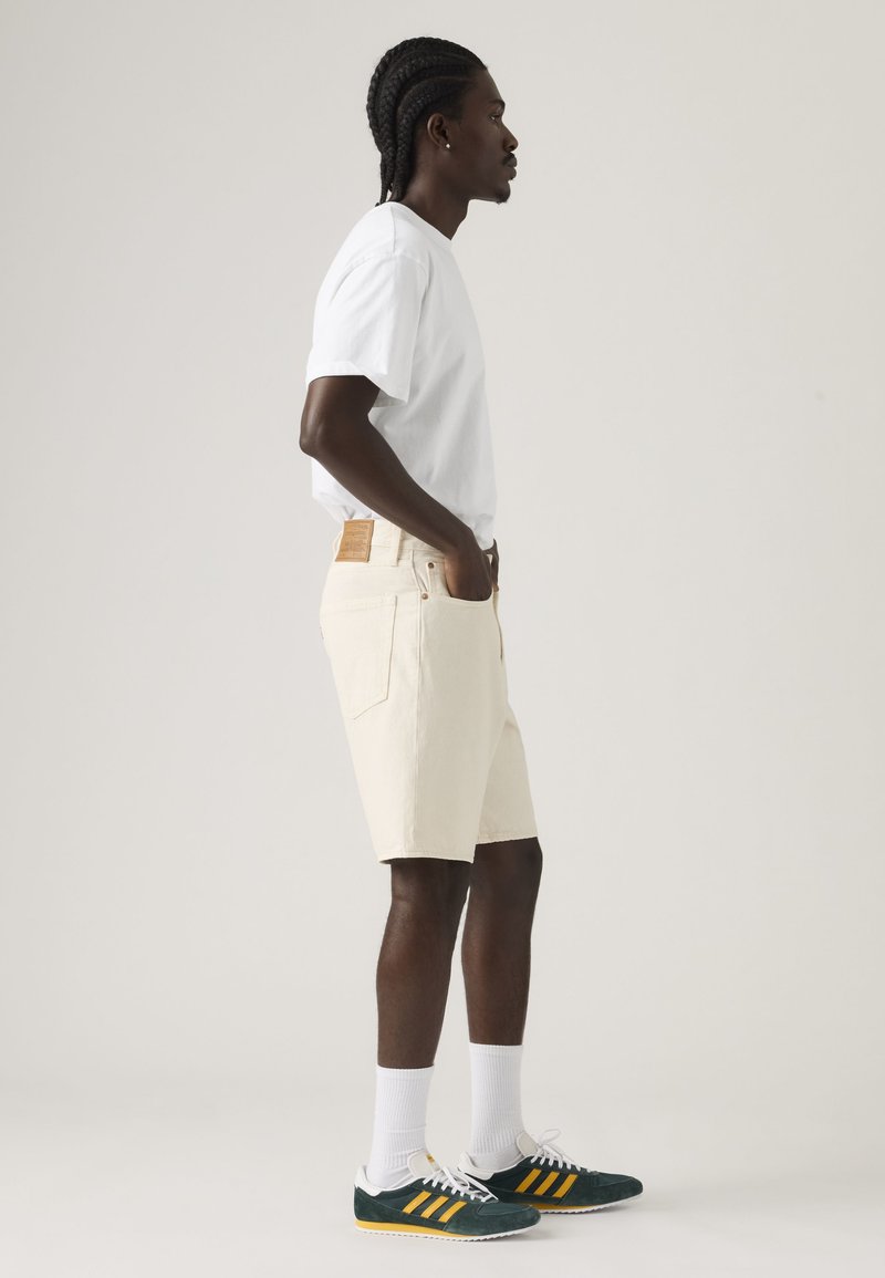 Uomo con capelli intrecciati che indossa una maglietta bianca, pantaloncini di jeans color crema, calzini bianchi e scarpe da ginnastica verde scuro con strisce gialle, in piedi di profilo.