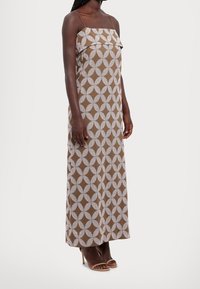 Brown maxi dress with white geometric pattern, strapless design, textured ruffle at the top, and a straight silhouette. Heels visible below.