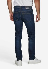 Dunkelblaue Jeans aus Denim im Slim-Fit, mit Gesäßtaschen, hellen Nähten und einem schwarzen Markenlabel am Bund. Weiße Turnschuhe sind sichtbar.
