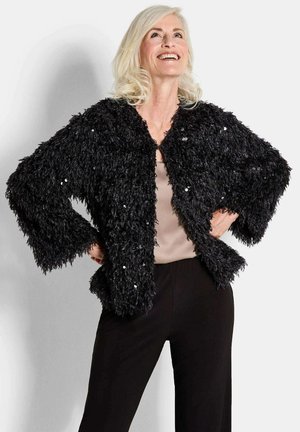 Veste noire texturée avec une finition duveteuse, à l'avant ouvert et aux manches trois-quarts, ornée d'accents en sequins. Portée par-dessus un top beige et un pantalon noir.