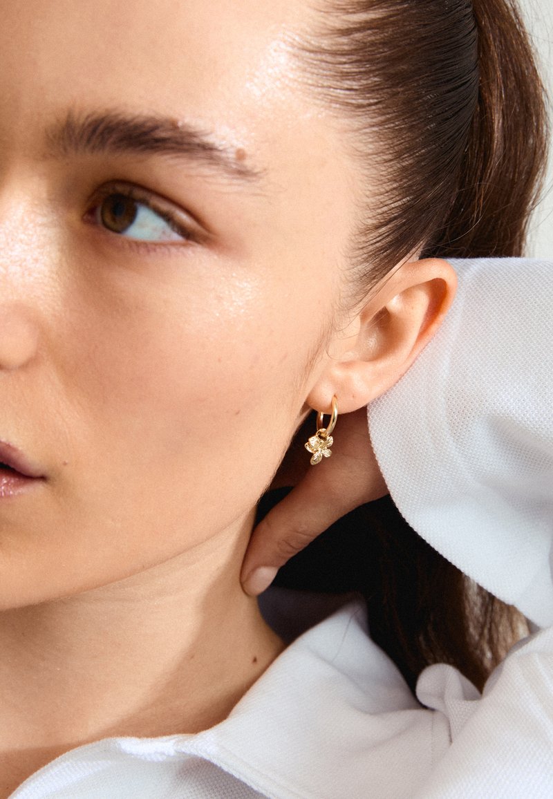 Boucles d'oreilles en or en forme de cerceau avec un charm floral contenant un petit gemme clair, portées par une personne à la peau claire et aux longs cheveux bruns.