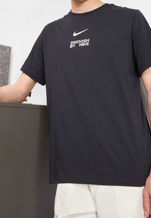 Persona que lleva una camiseta negra de Nike con texto blanco "Swoosh by Nike", de pie en el interior con un brazo apoyado en un armario.