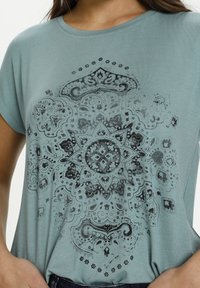 Světle tyrkysové tričko s krátkými rukávy, které má na středu detailní černý mandala potisk. Měkká tkanina s volným střihem.