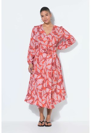 Frau steht mit der Hand in der Hüfte, trägt ein langärmeliges, mittelanges Kleid mit orange-pinkem Blattmuster und schwarze Sandalen vor weißem Hintergrund.