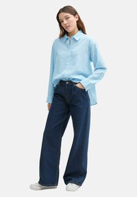 Camicia button-up a righe blu chiaro con maniche lunghe, abbinata a jeans a gamba larga blu scuro e sneakers bianche. Outfit semplice e casual.