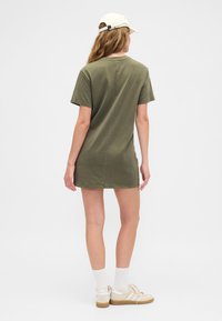 Olíva zöld pamut pólóruha rövid ujjakkal, kerek nyakkivágással és egyenes szabással, fehér zoknival és tornacipővel párosítva.