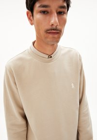 Beige Sweatshirt mit hohem Kragen und langen Ärmeln. Verfügt über ein kleines, weißes Logo auf der Brust und besteht aus strukturiertem Baumwollstoff.