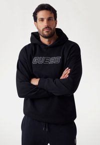 Felpa nera con logo ricamato "GUESS", maniche lunghe, tasca a marsupio frontale, realizzata in materiale morbido, abbinata a pantaloni neri coordinati.
