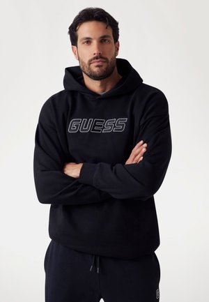 Schwarzes Hoodie mit gesticktem "GUESS"-Logo, langen Ärmeln, vorderer Kängurutasche, aus weichem Material, kombiniert mit passenden schwarzen Hosen.