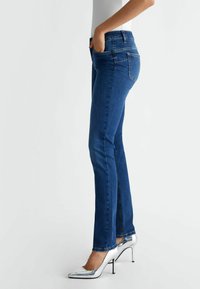 Jeans in denim blu con una forma aderente, caratterizzati da tasche frontali, un fondo leggermente svasato e dettagli cuciti. Indossati con tacchi alti argento.
