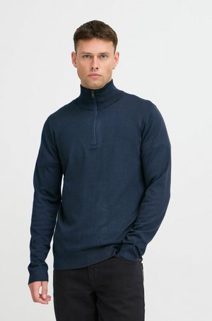 Pullover blu navy con colletto alto e zip parziale, realizzato in tessuto testurizzato. Maniche lunghe e polsini aderenti, abbinato a pantaloni scuri.