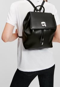 Musta nahkainen reppu, jossa on läppäkiinnitys, hopeinen logo-aksentti, säädettävät hihnat ja sileä pinta.