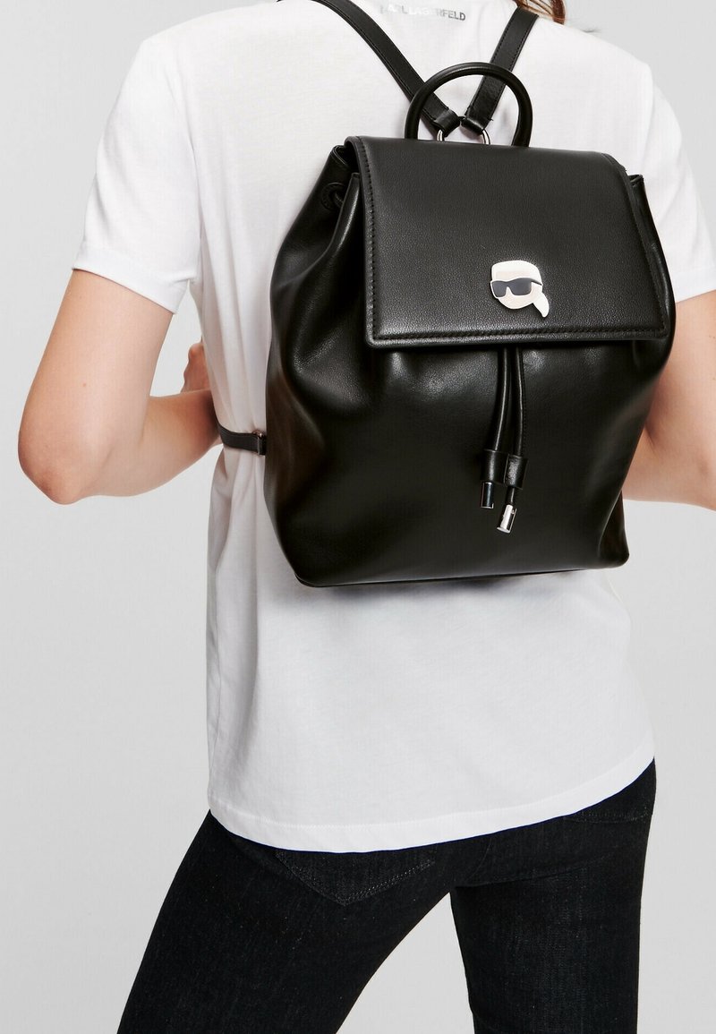 Musta nahkainen reppu, jossa on läppäkiinnitys, hopeinen logo-aksentti, säädettävät hihnat ja sileä pinta.