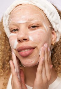 Persoon met krullend haar en een witte haarband die schuimende reiniger op hun gezicht aanbrengt, waardoor een gladde huidstructuur en subtiele plekjes zichtbaar worden.