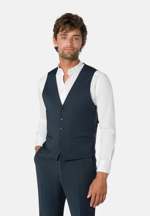 Mann trägt eine dunkelblaue formelle Weste, passende Hose und ein weißes Hemd mit Knöpfen, steht mit einer Hand in der Tasche vor einem schlichten Hintergrund.