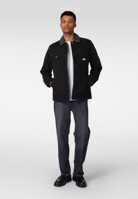 Svart lerretsjakke med kordfløyelskrage, to frontlommer og messingknapper med trykk. Kombinert med mørke denimjeans og svarte sko.