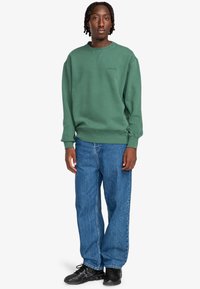 Grüner Baumwollpullover mit gerippten Bündchen und Saum, ausgestattet mit einem dezenten Logo. Kombiniert mit weiten blauen Denim-Jeans und schwarzen Sneakern.