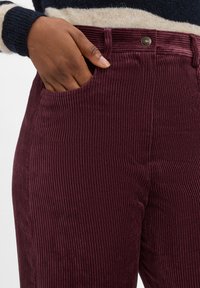 Pantalons en velours côtelé bordeaux avec des côtes verticales, dotés d'une poche latérale et d'un détail en métal sur la taille. Texture douce.