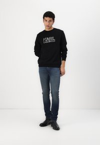 Fekete pulóver "KARL LAGERFELD" logóval, textúrált nyomattal, kék, slim-fit farmerral és fekete cipővel párosítva, fehér háttér előtt.